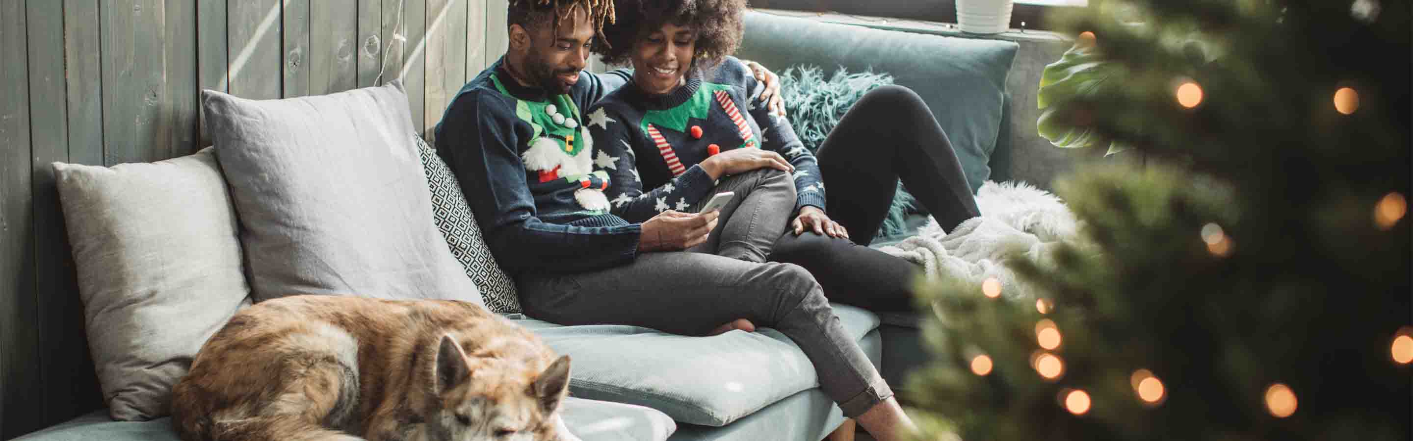 A man and woman lounge on their couch with their dog in front of a Christmas tree